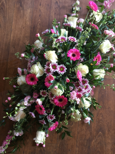 Blushing Memories Casket Spray - This beautifully feminine casket spray is a delicate and uplifting tribute, featuring a romantic mix of soft cream roses, hot pink gerberas, pink lisianthus, and bi-color chrysanthemums. Accented with light pink stock flowers and lush greenery, the arrangement exudes warmth, love, and grace. The varying shades of pink paired with creamy whites create a gentle yet vibrant visual that celebrates a life full of kindness and joy. Perfect for honoring a beloved individual with a touch of elegance, tenderness, and heartfelt remembrance.